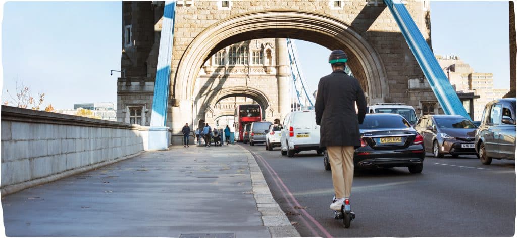 An image of a man riding away on a Dott over a bridge. A classic bright red London bus is a few cars ahead of him. The sun reflects on the bridge.
