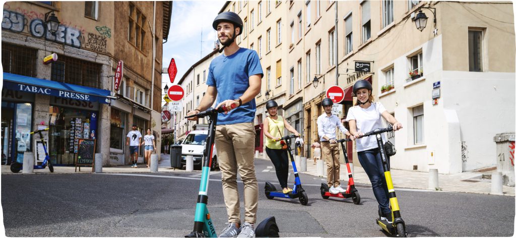 A group of four people riding scooters coming around a corner in a sunny European city. All are wearing helmets and each has a different colored scooter, one red, yellow, green and blue.