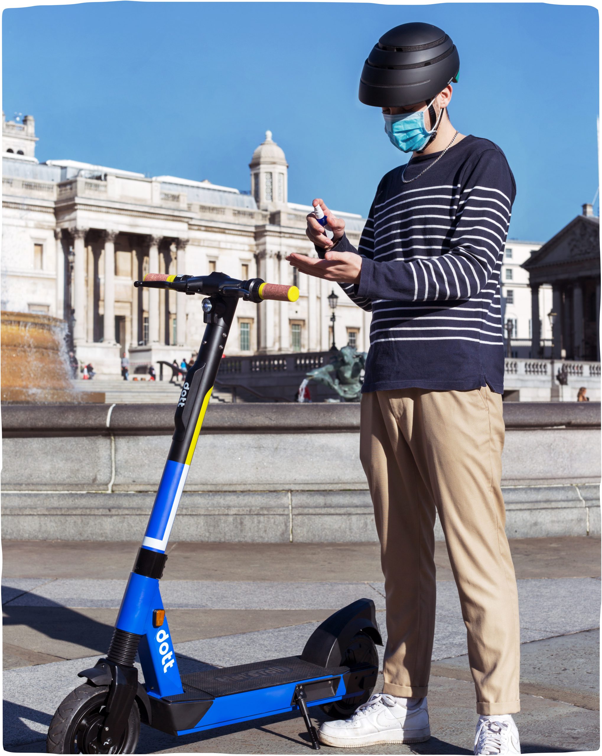 A young man stands next to a blue Dott scooter wearing a helmet and face mask. He is spraying his hands with disinfectant.