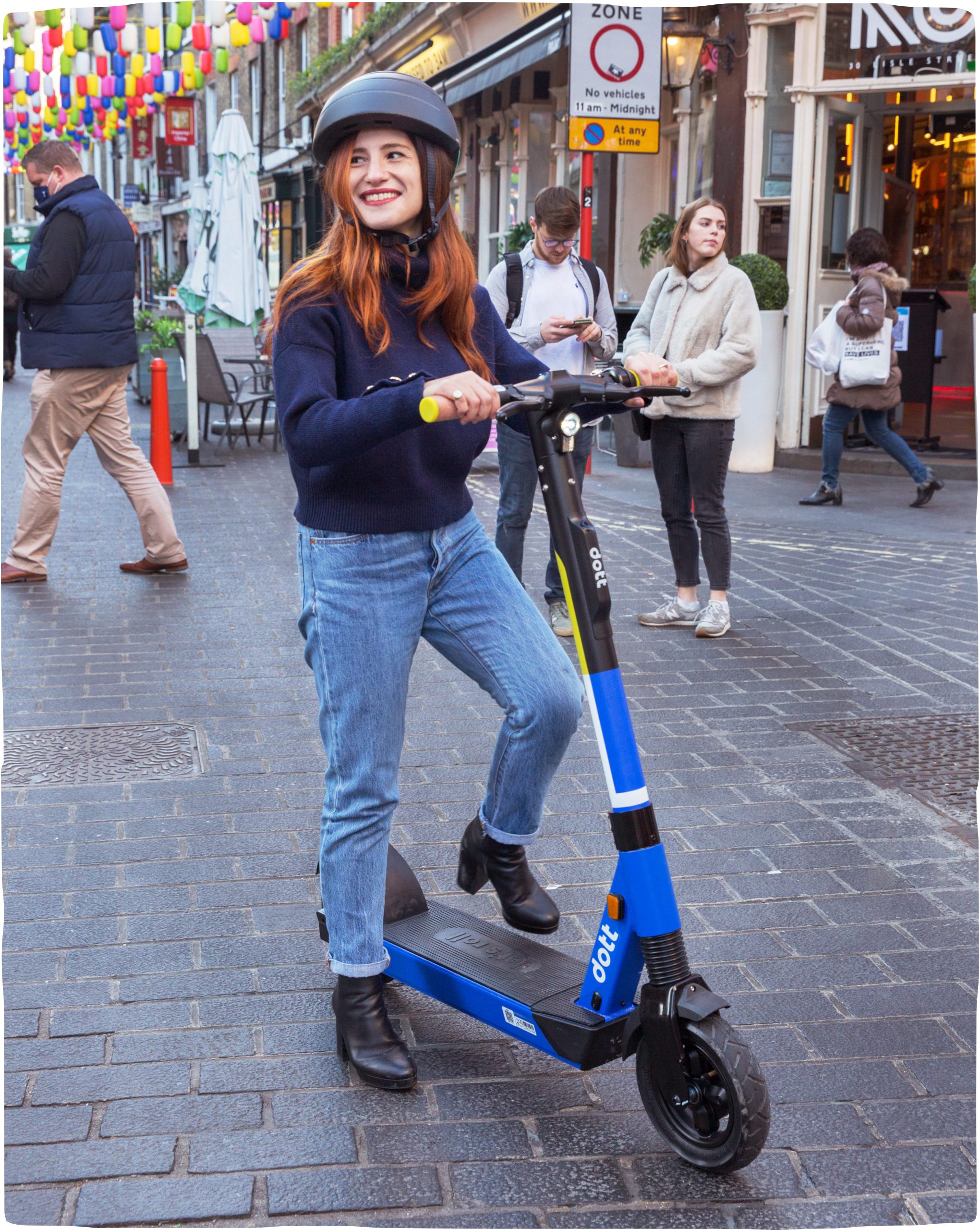 A woman steps onto a blue Dott scooter, smiling and looking into the distance. She is wearing a helmet.