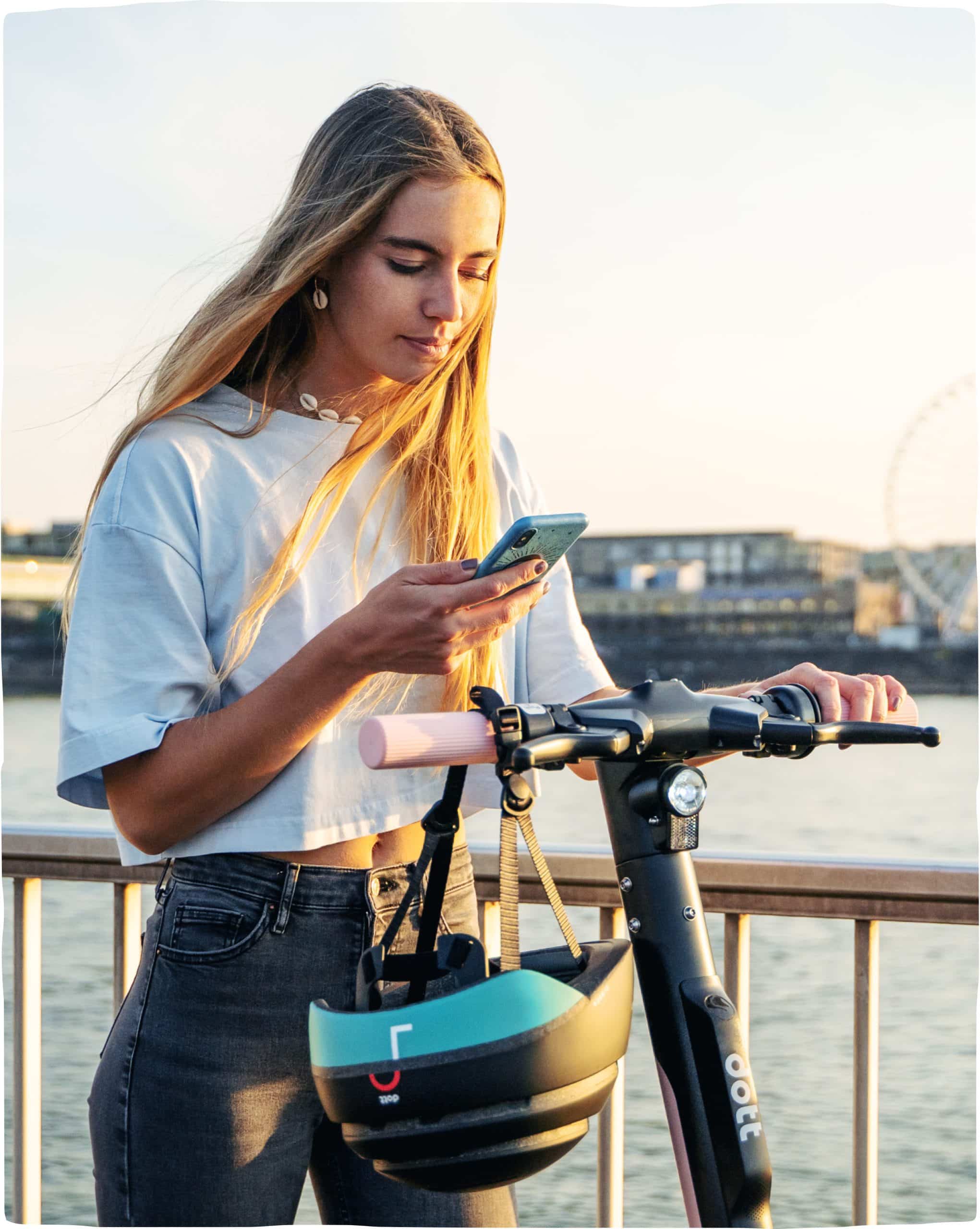 A young woman stands on a Dott and checks her phone in her right hand while she holds the scooter steady with her left hand. The evening sunlight reflects golden on her blonde hair.