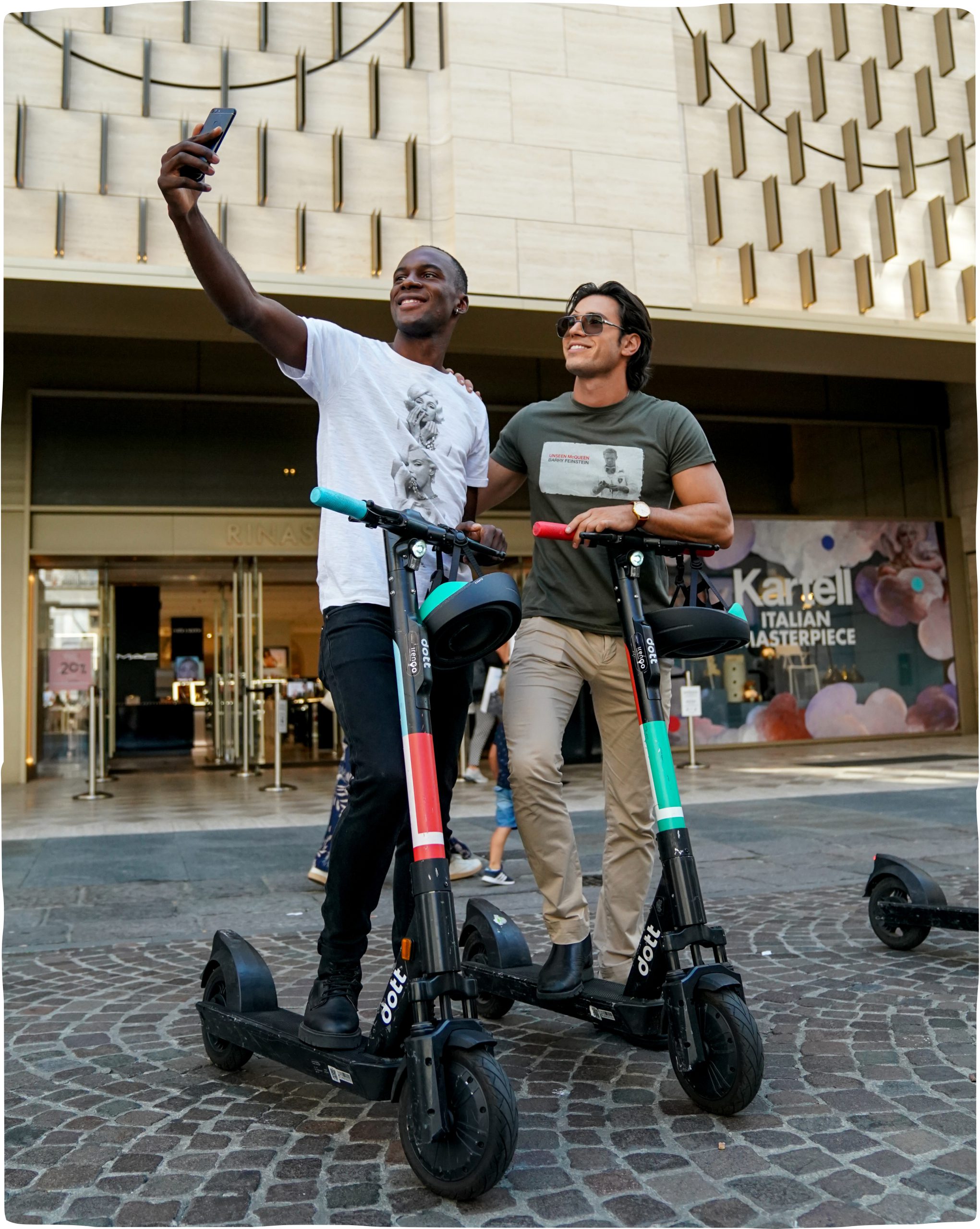 Two men take a selfie together on Dotts on a cobblestone city street.  One man holds the phone with his arm outstretched as the other casually rests his foot on the scooter.