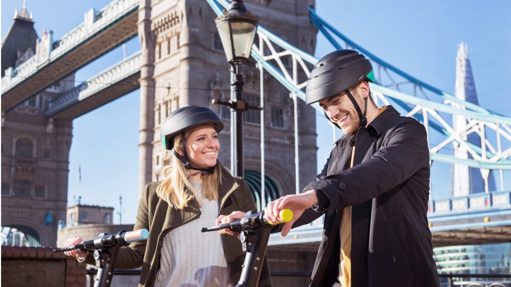 A man and a woman standing together on a sunny day smiling at each other with two e-scooters, with the London Bridge in the background.