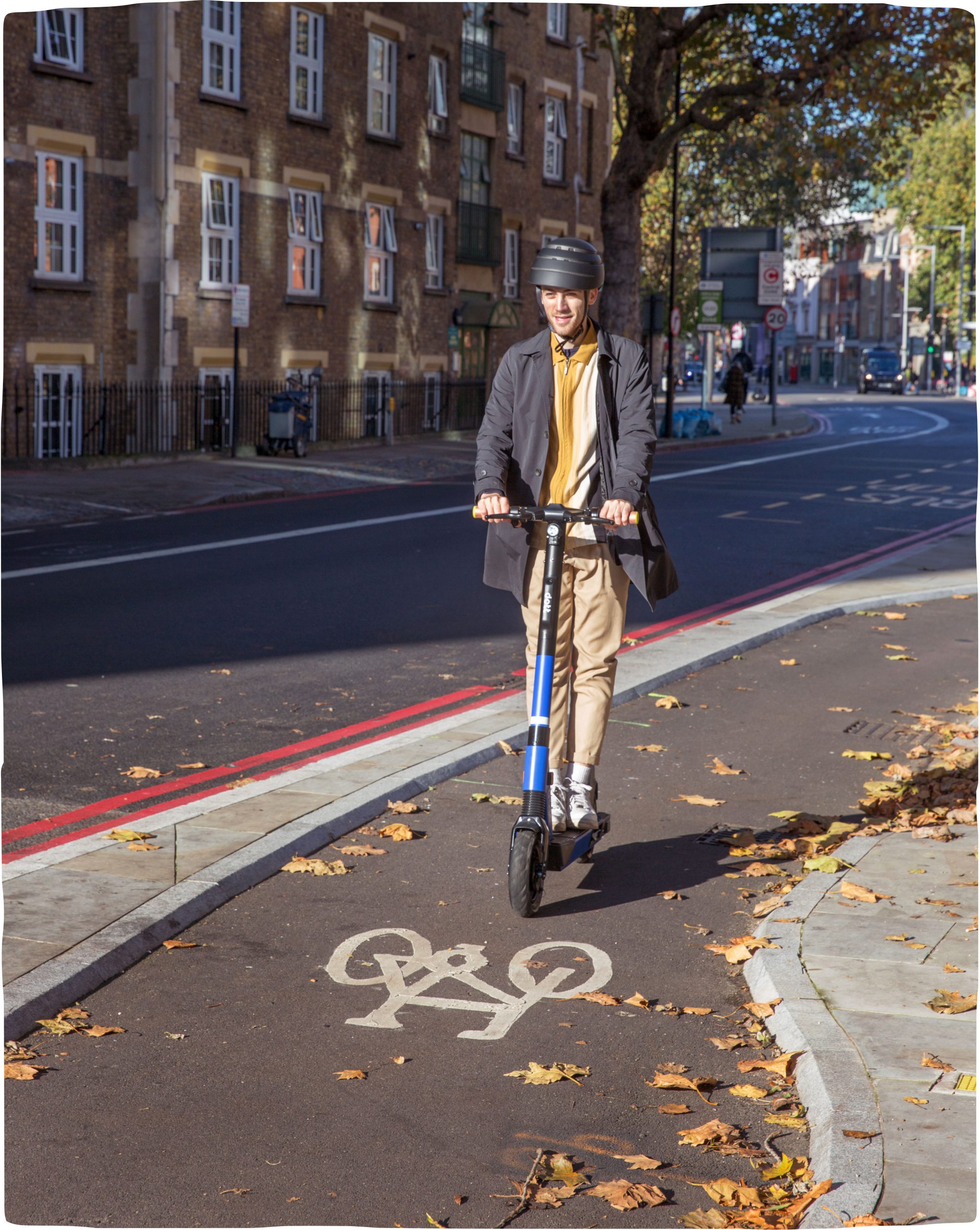 A man wearing a helmet and riding an e-scooter in a bicycle lane on a sunny autumn day in London.