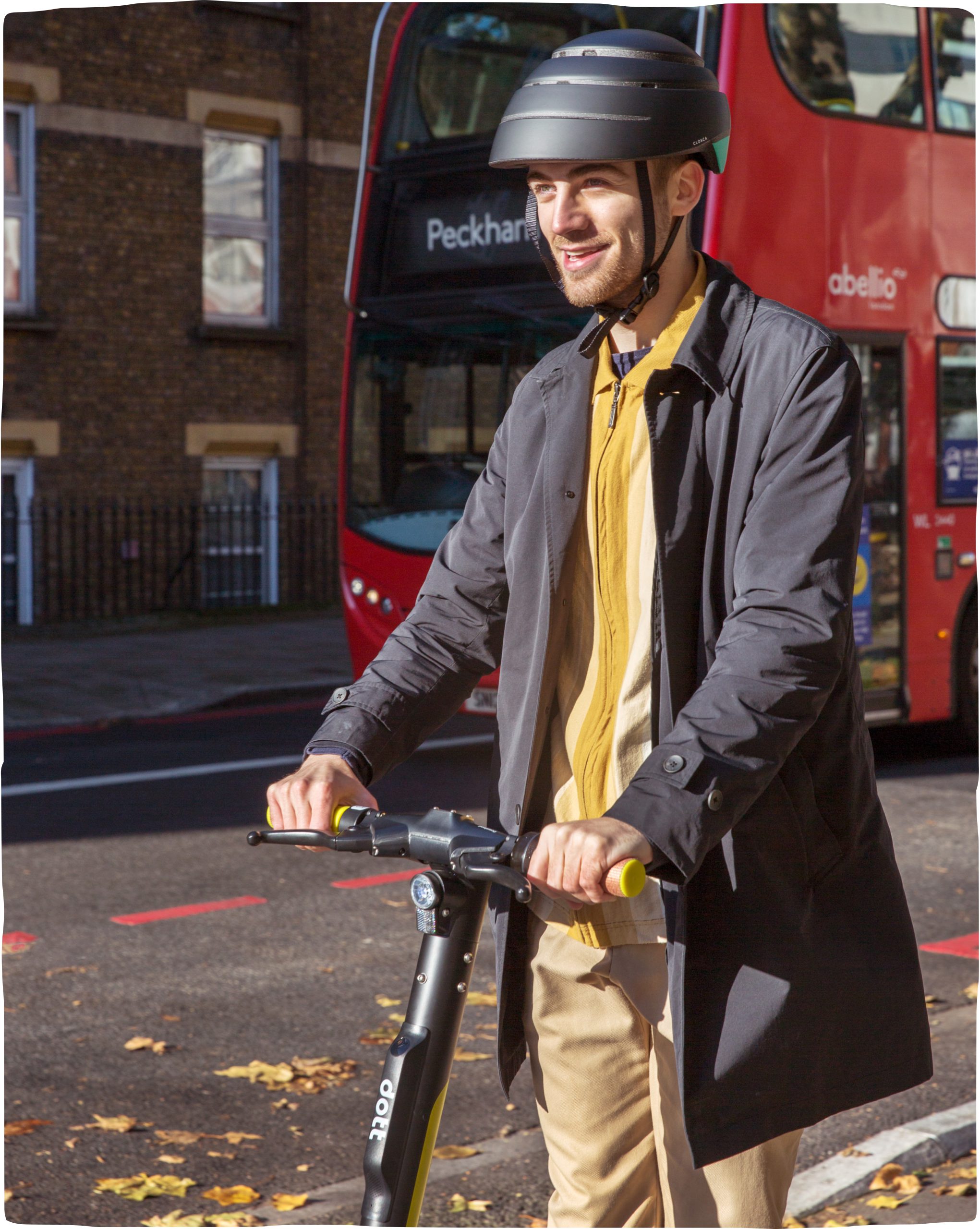 A man wearing a helmet and riding an e-scooter down a London street with a red double-decker bus in the background.