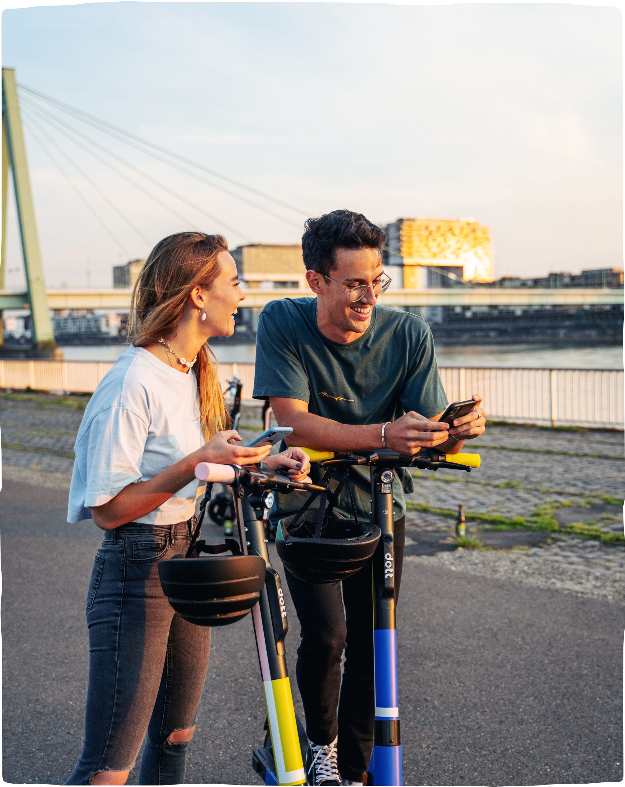 A woman and a man smiling and looking at their phones while they take a break from riding scooters, with a bridge and building in the sun behind.