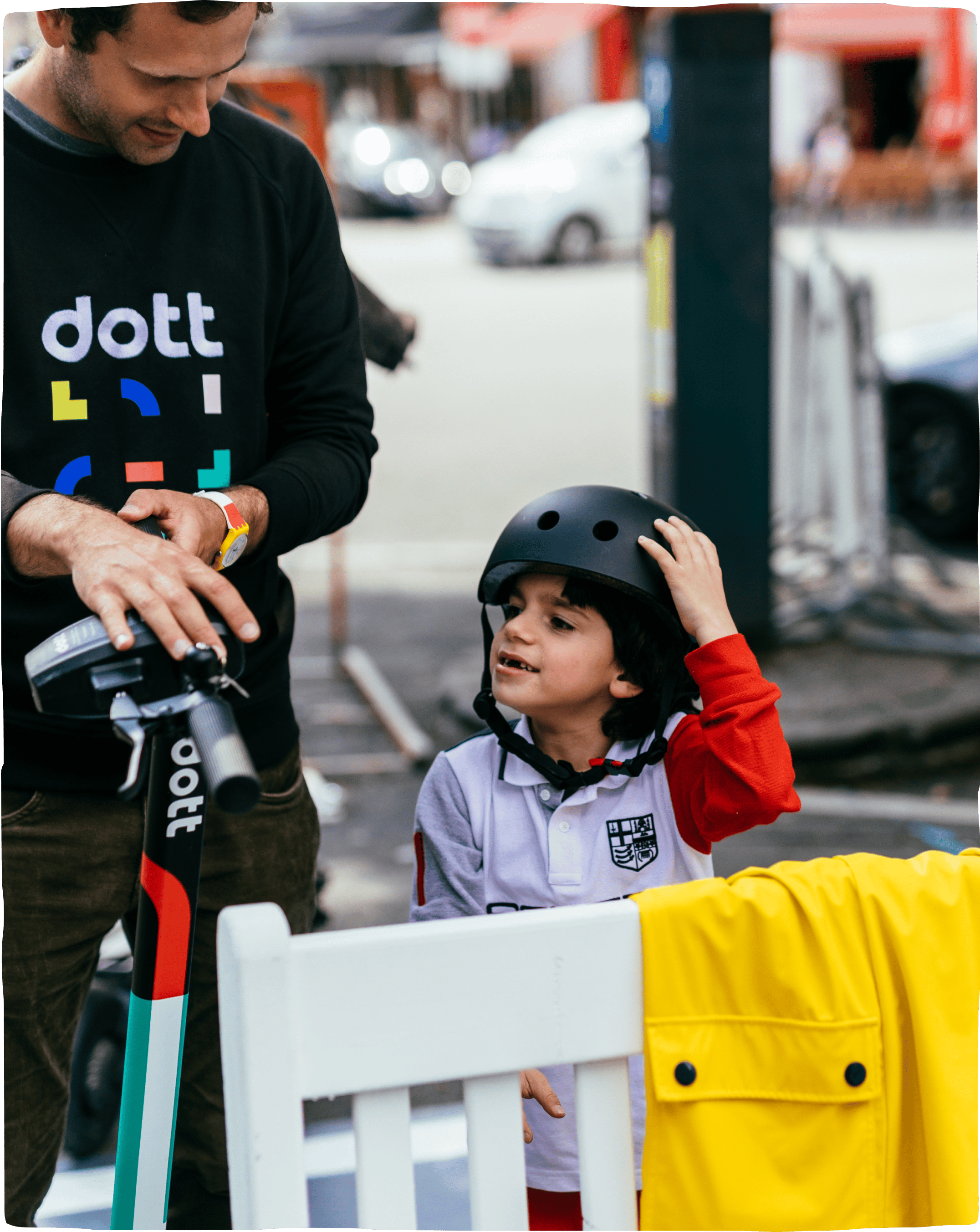 A man in a Dott branded sweatshirt standing with a young boy, showing him the e-scooter and helping him test out a helmet.