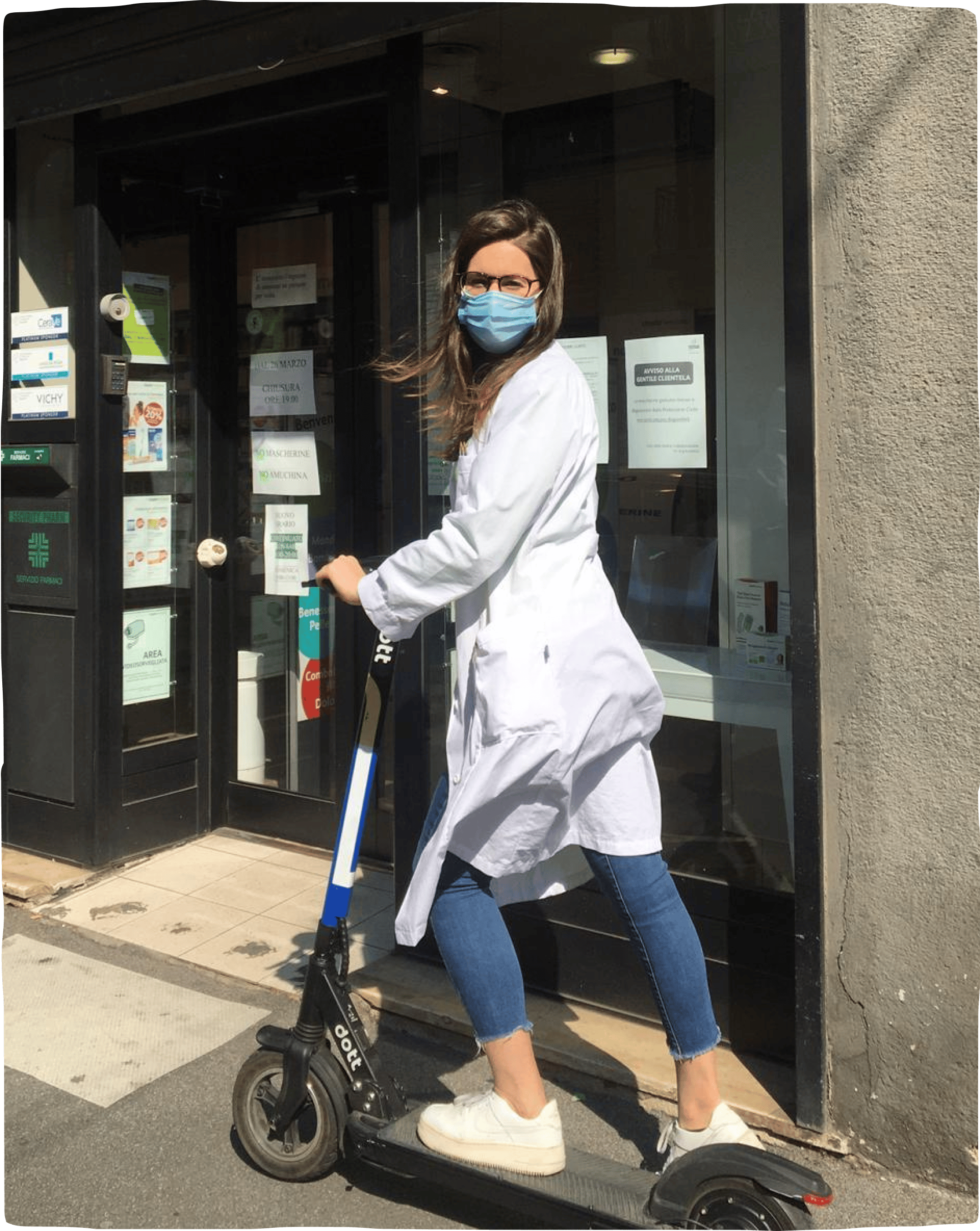 An A&E worker wearing a mask and white coat, posing with a Dott e-scooter outside of a pharmacy.