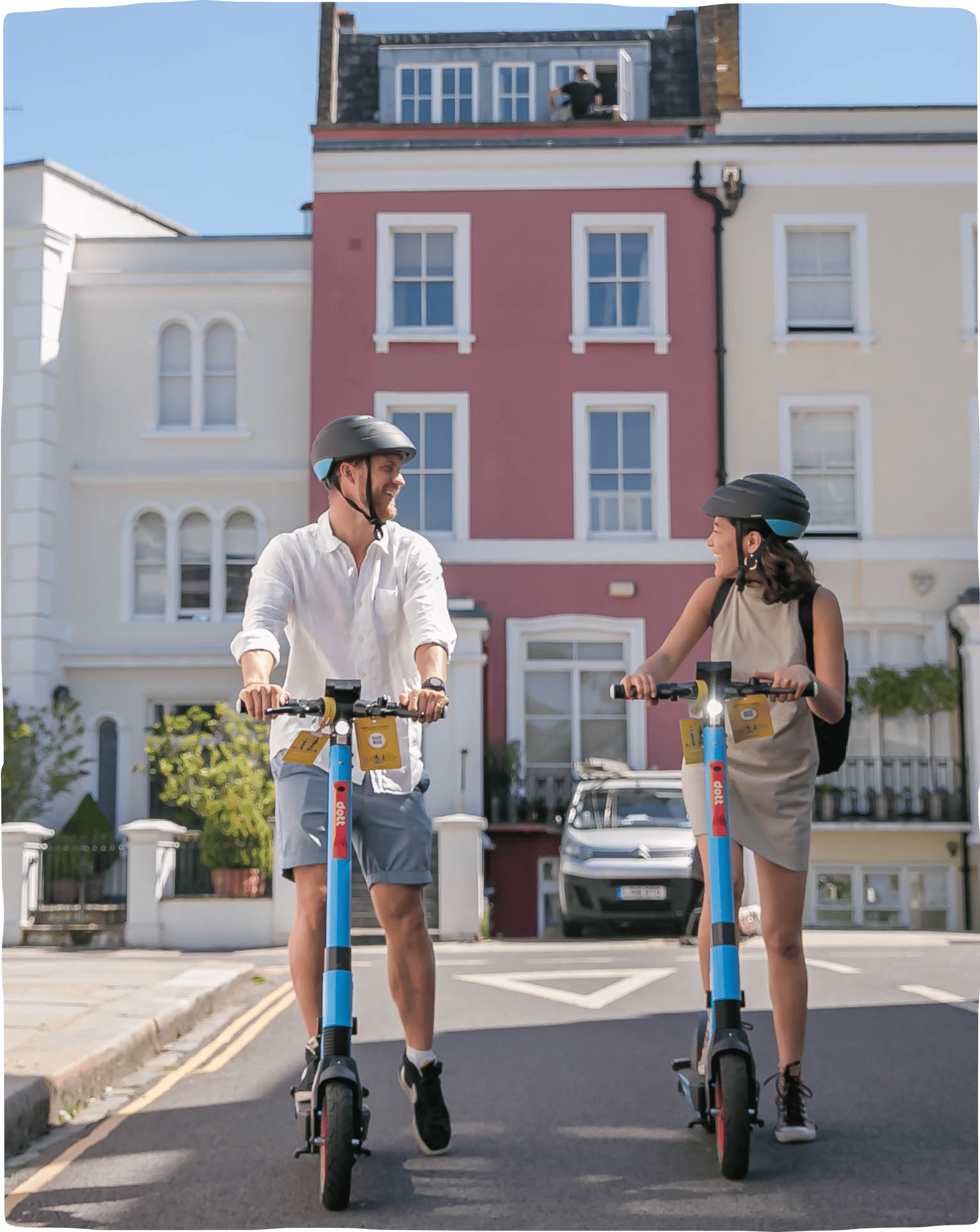 A man and a woman wearing helmets and smiling at each other while riding down a sunny street, with row houses in the background.