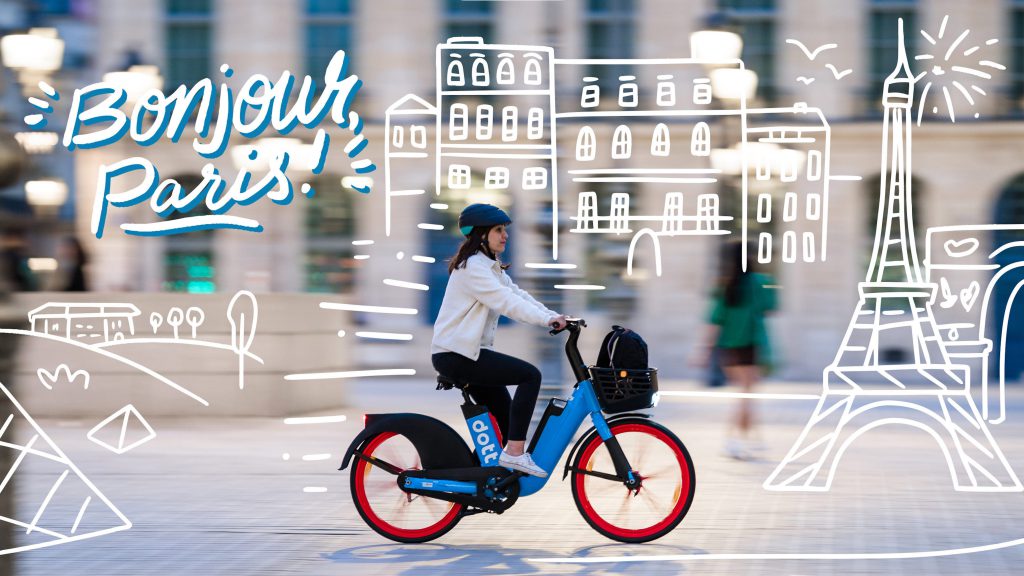A header image of a woman riding a blue Dott e-bike. The sun reflects on the sidewalk. The background is blurred to indicate movement, and there are white illustrations of a cityscape in the background