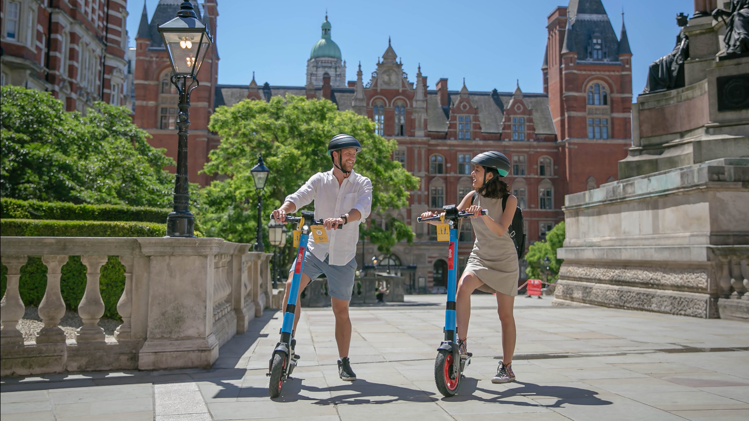 A man and a woman wearing helmets stand smiling and casually holding their Dott scooters while they talk to each other. The sun shines on the grand buildings behind them.