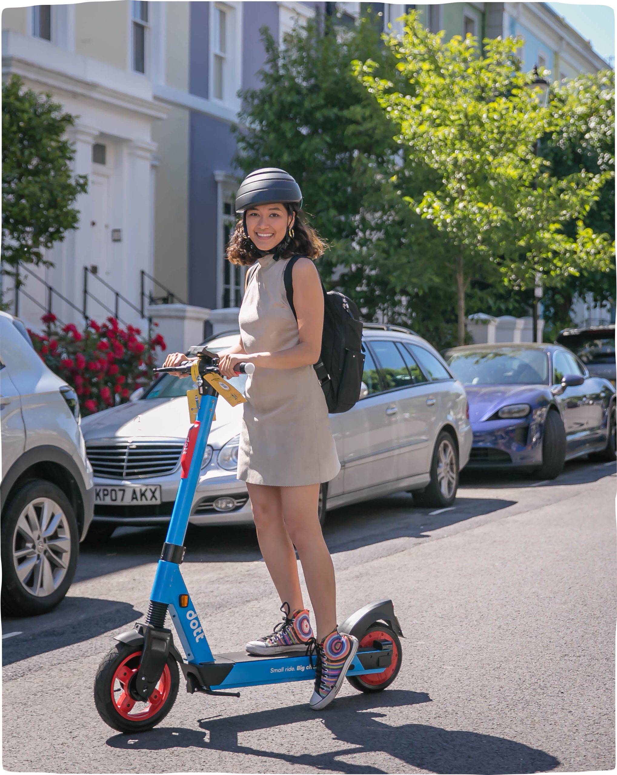An image of a young woman holding the handlebars of her blue Dott scooter and smiling towards the camera. The sun shines on the tree behind her.
