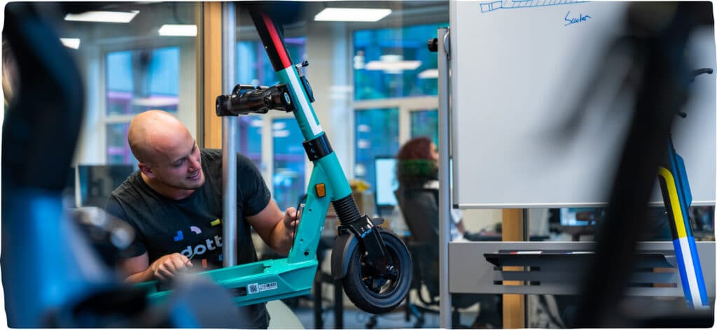 A Dott employee from the Hardware team working on an e-scooter in the workshop with the office in the background.