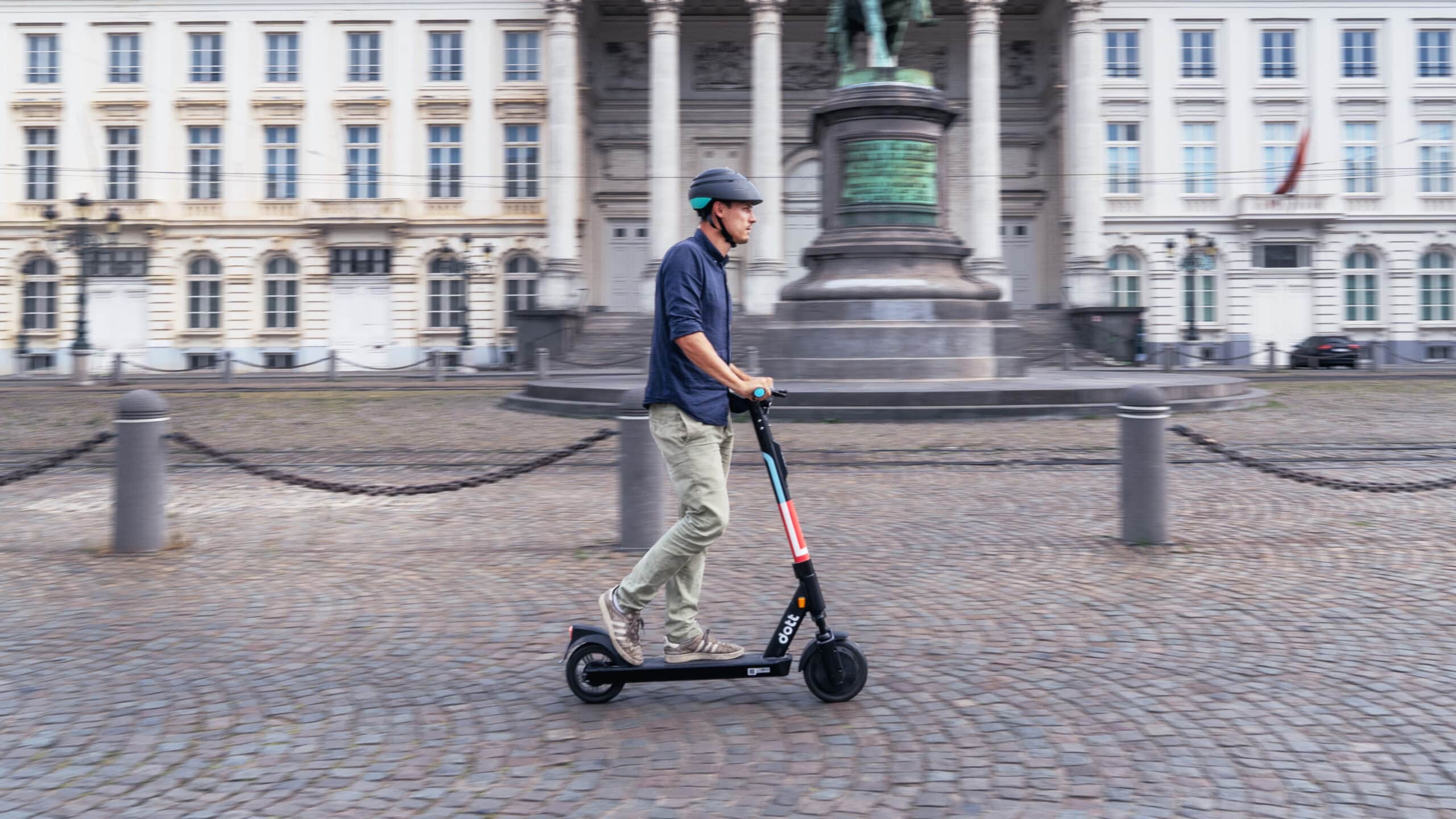A man riding a scooter with a helmet through a European historic landscape with cobblestone streets.