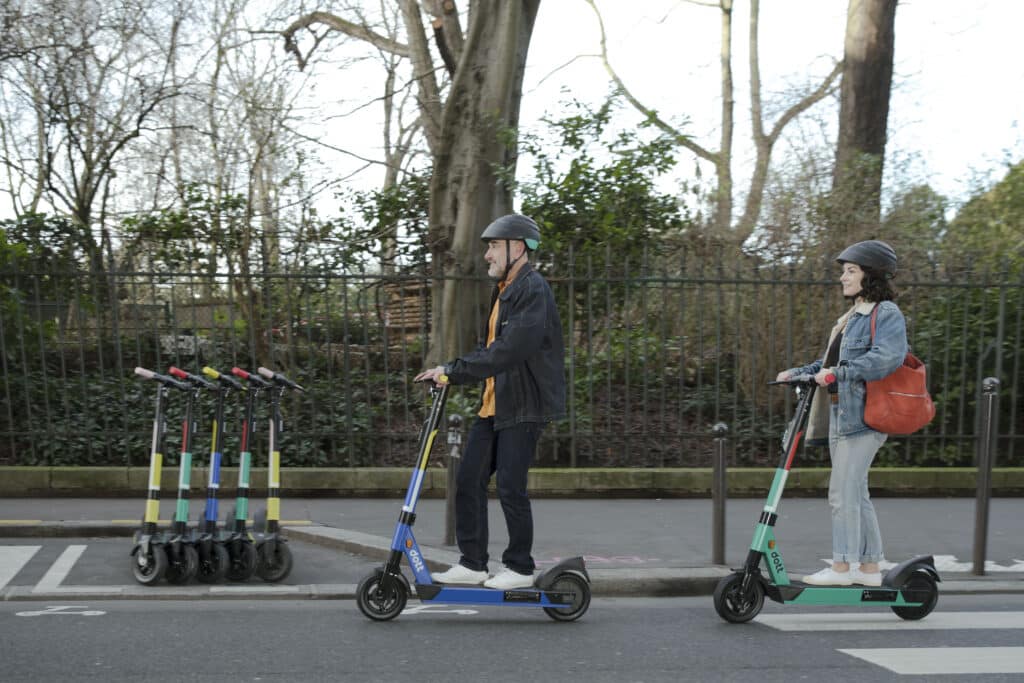 A man and a woman ride Dott scooters with helmets, smiling. The man rides in front of the woman and the background is blurry to show they are moving.