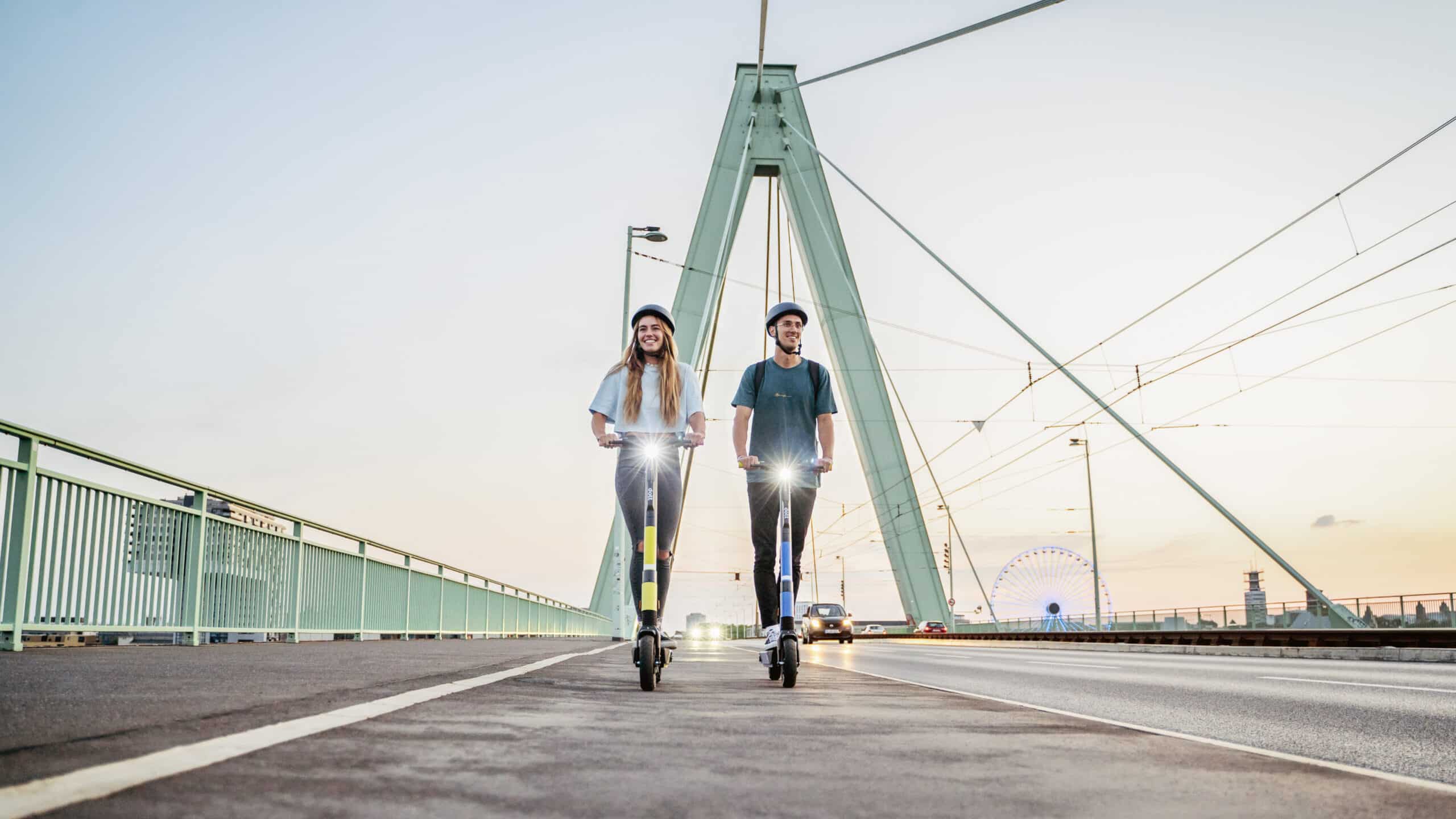 Een foto van een vrouw en een man die naast elkaar op een brug op Dott-scooters rijden.
