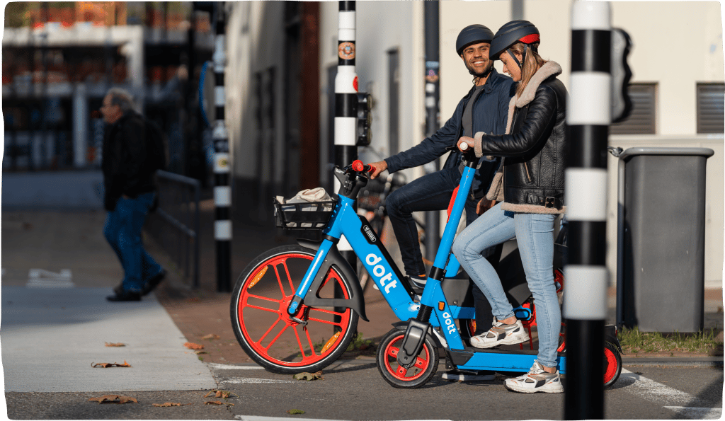 A photo of a man and woman riding blue Dott e-bikes and e-scooters in a city, stopped at a stoplight and smiling at each other.