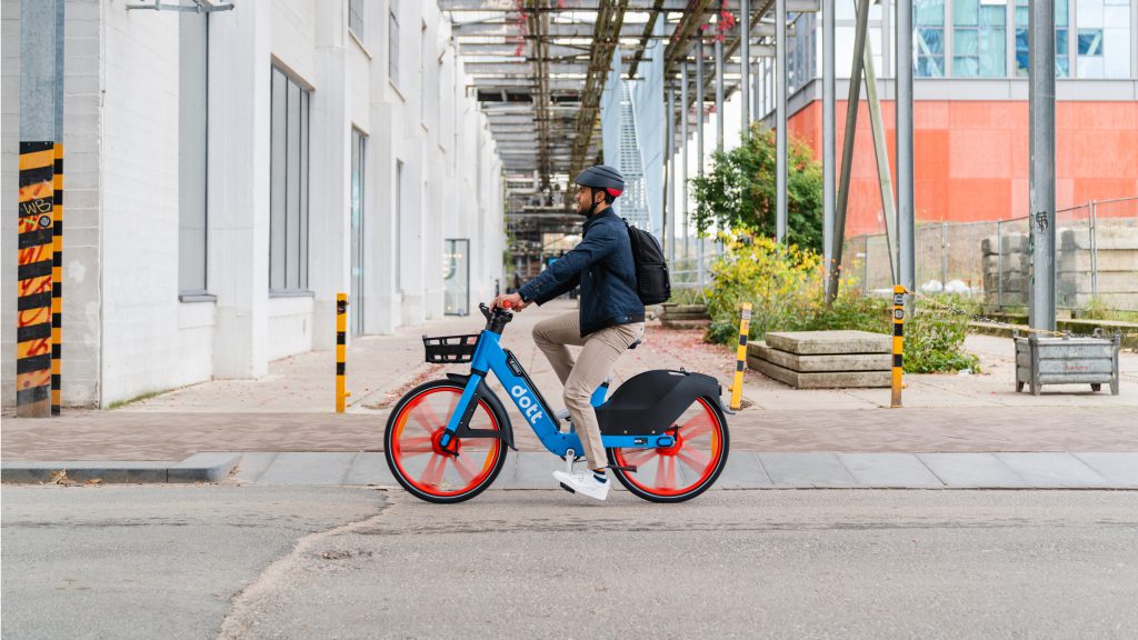 An image of a man riding a blue Dott e-bike with an industrial background.