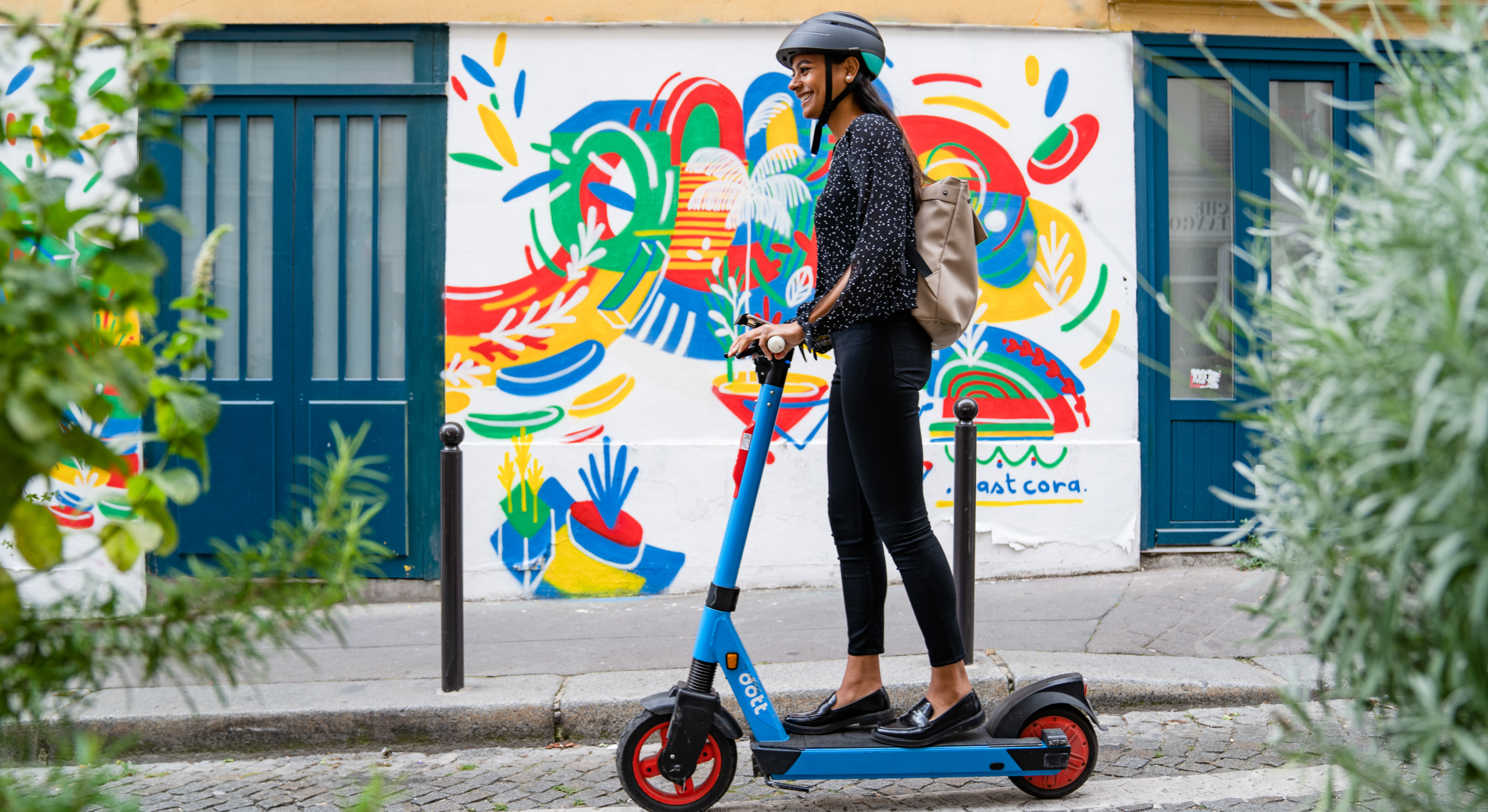 An image of a woman riding a blue Dott e-scooter in a city, with a colorful street mural in the background.