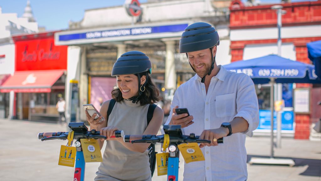 A young man and woman stand next to each other, wearing helmets and smiling  down at their phones. They hold the Dott e-scooter handlebars with their left hands. Bright yellow illustrated hangtags hang from both scooters.