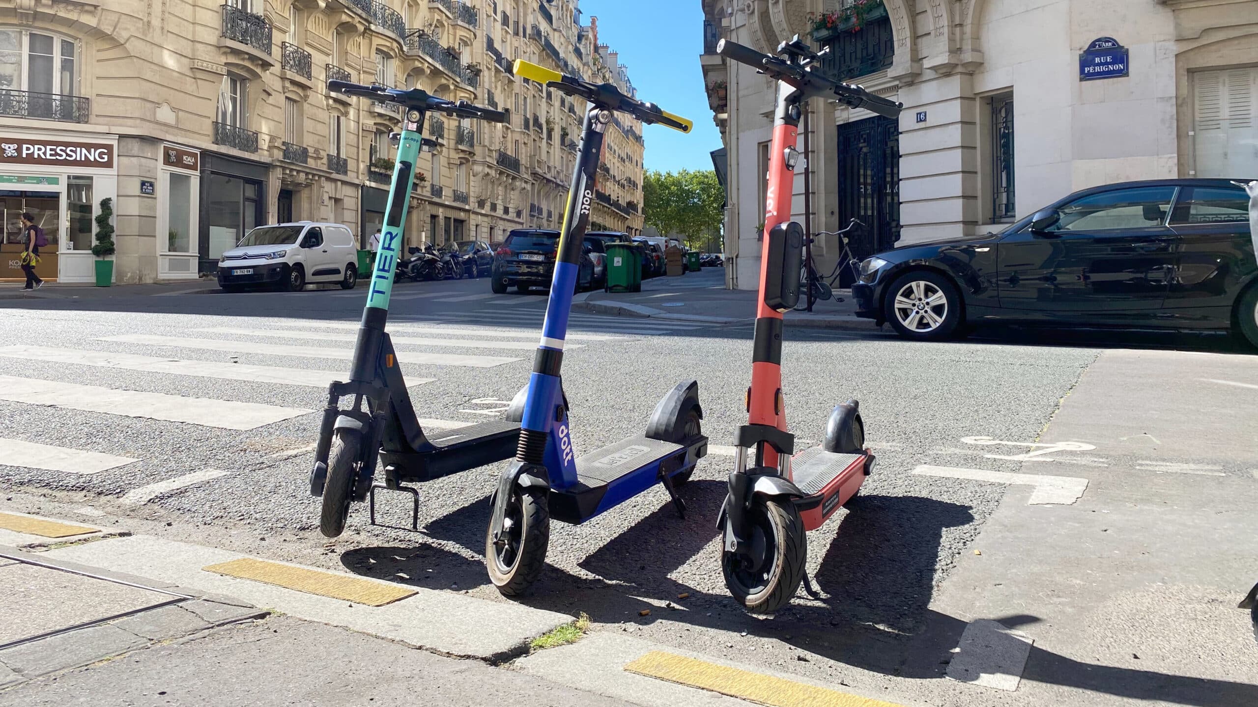 A Tier e-scooter, a Dott e-scooter and a Voi e-scooter are all parked in a line on a street in France, the weather is sunny.
